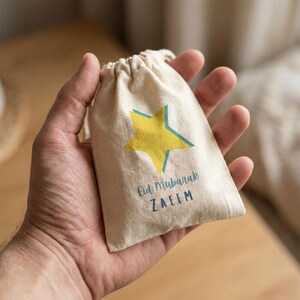 Può includere: Un piccolo sacchetto beige con coulisse, con una stella gialla e dettagli turchesi. La borsa ha la scritta "Eid Mubarak" e il nome "ZAEEM" stampati in turchese. La borsa è tenuta in una mano.