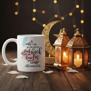 Peut inclure: Tasse en céramique blanche avec une anse, ornée de l'inscription "The only time you should ever look back is to see how far you've come Alhamdulilah". La tasse est décorée d'accents aquarelle bleus et roses. Lanternes décoratives et croissant de lune en arrière-plan.