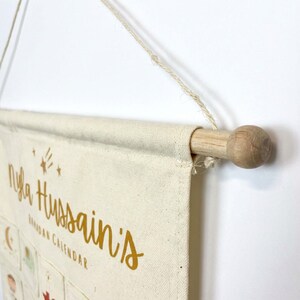 May include: Cream-colored Ramadan calendar with gold lettering that reads "Nyla Hussain's Ramadan Calendar." The calendar is suspended by a wooden dowel and a natural fiber string. The design includes star and crescent moon graphics.
