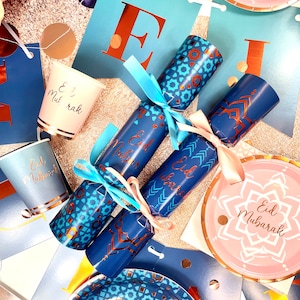 Op de afbeelding: Blauwe en gouden feestbenodigdheden met een feestelijk Eid Mubarak-thema. De afbeelding toont papieren bekers, borden en feestcrackers met een blauw en gouden geometrisch patroon. De tekst "Eid Mubarak" staat op de borden en bekers.