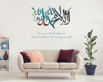 Op de afbeelding: Een witte muur met een grote teal en blauwe islamitische kalligrafie muursticker. De sticker bevat de zin "Er is geen god dan Allah en Mohammed (saw) is de boodschapper van Allah". De sticker is geplakt op een witte muur in een woonkamer met een bank en een plant.