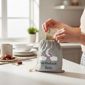 Può includere: Un sacchetto grigio con coulisse con un design arcobaleno e luna, e il testo "Eid Mubarak Isla". Una persona sta mettendo dei soldi nel sacchetto. Il sacchetto è su una superficie bianca.