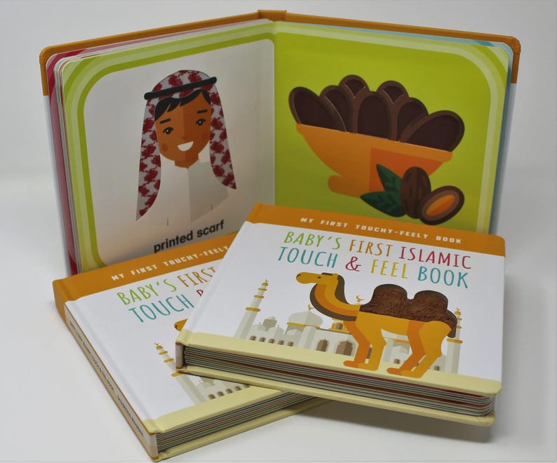 May include: A children's book titled "Baby's First Islamic Touch & Feel Book" with illustrations of a camel and a mosque. The open book shows images of a person wearing a printed scarf and dates in a bowl. The book has a yellow and white cover.