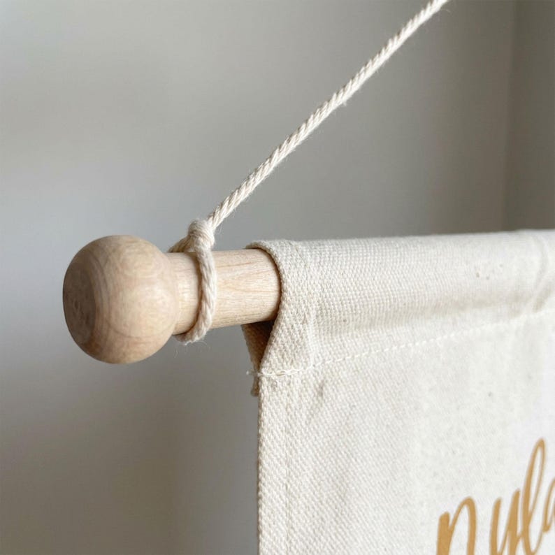 May include: A close-up of a beige canvas banner with a wooden dowel and a natural rope for hanging. The banner has gold lettering. The wooden dowel has a rounded end. The rope is tied around the dowel.