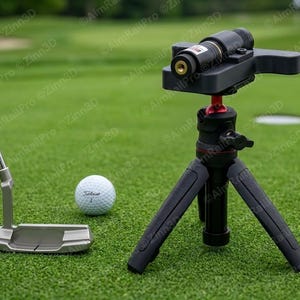 May include: A silver putter, a white Titleist 1 golf ball, and a black laser alignment device mounted on a small black tripod with a red adjustment knob. The laser device features a cylindrical barrel with a gold-colored aperture at the front.