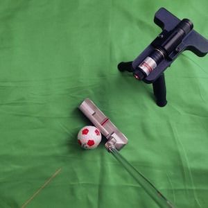 May include: A white and red golf ball sits on a green surface next to a silver putter with a red stripe. A black laser pointer on a tripod is in the top right corner of the image.