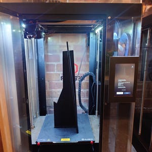 Peut inclure: Une imprimante 3D noire avec un boîtier en plastique transparent. L'imprimante imprime un objet noir sur une plateforme noire. L'imprimante a un bouton rouge sur le devant.