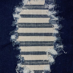 Puede incluir: Primer plano de una tela vaquera azul con un agujero que revela una tela a rayas blancas y azules debajo. El agujero tiene un borde deshilachado.