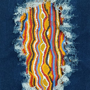 May include: A close-up of a denim fabric with a large hole revealing a colourful patterned fabric underneath. The pattern is made up of stripes and circles in shades of red, orange, yellow, blue, and white.