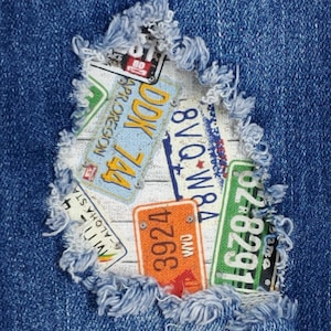 May include: A close-up of a ripped denim fabric with a patch of various license plates sewn on. The license plates are from different states and have various numbers and letters on them. The license plates are in a variety of colors, including red, blue, green, and yellow.