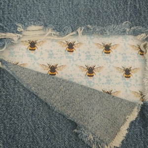 Może przedstawiać: Zbliżenie na parę niebieskich dżinsów z przetartym fragmentem, który odsłania białą tkaninę z powtarzającym się wzorem żółtych i czarnych trzmieli.
