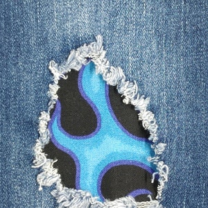 Op de afbeelding: Blauwe denim jeans met een gescheurd gat dat een zwarte stoffen patch met een blauw en paars vlammenpatroon onthult.