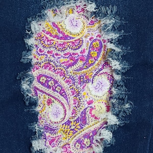 Puede incluir: Primer plano de un parche de mezclilla desgastada con un vibrante diseño de cachemira. El parche es una mezcla de morado, rosa, amarillo y blanco, con un borde deshilachado, agregando un toque único a la mezclilla.
