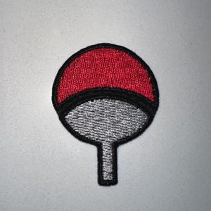 May include: Embroidered patch featuring a red and black design with a silver-gray lower section and a stem. The patch has a circular top with a black border and a stem-like base. The patch is likely for clothing or accessories.