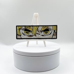 May include: Embroidered patch featuring a detailed depiction of anime-style eyes. The eyes are yellow with black outlines, set against a white background, and framed by a black border. The patch is rectangular and appears to be on a white display stand.