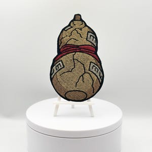 Peut inclure: Écusson brodé représentant une gourde avec une bande rouge et du texte. Le patch a un fond beige avec des coutures noires et une texture craquelée. Il est présenté sur un support blanc.