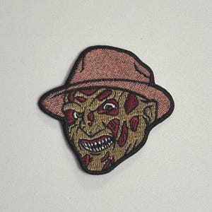 Peut inclure: Écusson brodé représentant le visage d'un personnage de film d'horreur. L'écusson présente un chapeau marron, un visage cicatrisé avec des détails rouges et un sourire menaçant. L'écusson est bordé de noir.