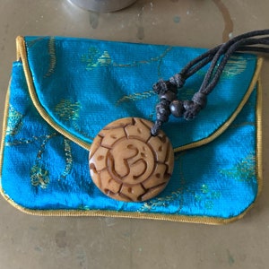 May include: A teal blue silk pouch with a gold trim and a wooden charm with a carved symbol. The pouch is on a brass tray with a lit candle and a burning incense cone.