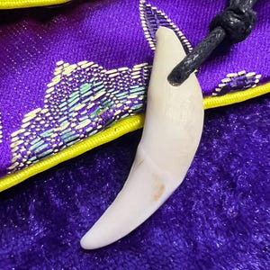 May include: A curved, off-white tooth pendant hangs from a black cord, resting on a small, purple satin pouch. The pouch features a vibrant floral pattern in red, green, and gold, with a bright yellow trim and a matching yellow snap button closure.