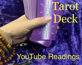 May include: A purple tarot deck with a triple moon and pentacle design is held in a hand. The words "Tarot Deck" and "YouTube Readings" are displayed in yellow text. A beaded bracelet is visible.