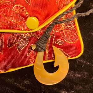 May include: A red pouch with a floral pattern and yellow trim, holding a beige fish hook pendant on a black cord. The pouch has a yellow button closure. The pendant is a light beige colour.