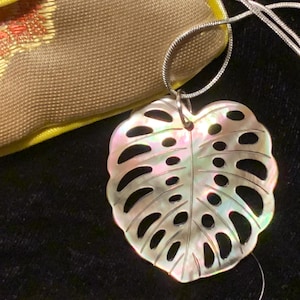 May include: A silver necklace with a Monstera leaf pendant. The pendant has a shimmering, pearlescent finish and multiple cut-out holes. The necklace is set against a dark backdrop, with a gold and beige pouch visible in the background.