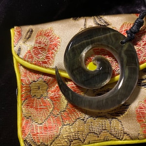 May include: A dark green, spiral-shaped pendant necklace with a black cord. The pendant rests on a floral patterned pouch with yellow trim. The background is black.