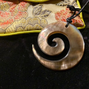 May include: A brown and black shell pendant with a spiral design hangs from a black cord. The pendant is on a yellow and gold patterned fabric background.