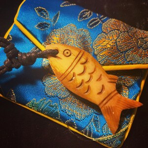 May include: A carved wooden fish pendant with a black cord, resting on a blue and gold patterned fabric pouch.