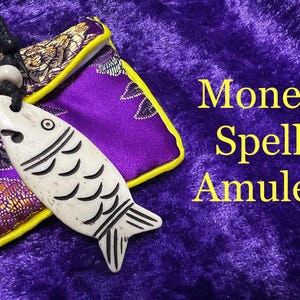 May include: A cream-colored fish-shaped amulet with black detailing hangs from a black cord. The amulet is set against a purple velvet background with the words "Money Spell Amulet" in yellow. A purple pouch with gold trim is also visible.