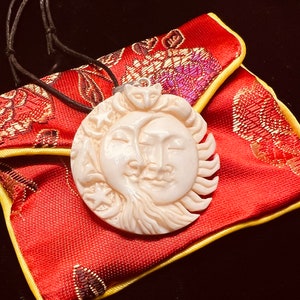 May include: A white carved bone pendant depicting a sun and moon with faces. The pendant is on a red and gold fabric pouch with a black cord.