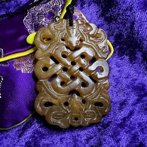 May include: A tan-colored, rectangular stone pendant featuring an intricate, openwork carved knot design. The pendant is suspended from a black cord and rests against a purple fabric pouch with a yellow trim and a floral pattern.