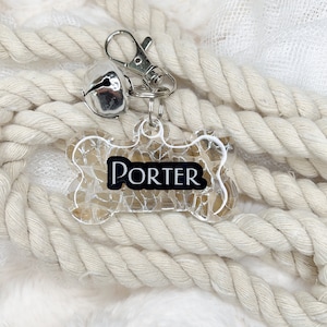 May include: A clear, bone-shaped dog tag with a silver bell and a silver clip. The tag has a cracked glass design and the name "PORTER" printed in black.