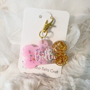 Op de afbeelding: Roze en gouden glitter hondenbot bedel met een gouden sluiting en de naam "Bella" erop. De bedel is op een witte kaart met de tekst "Deer Bella Craft".