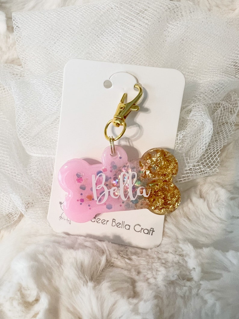 Op de afbeelding: Roze en gouden glitter hondenbot bedel met een gouden sluiting en de naam "Bella" erop. De bedel is op een witte kaart met de tekst "Deer Bella Craft".