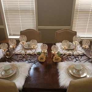 May include: Two gold metal candle holders with crystal accents and a white fur placemat on a wooden table. Each candle holder has five crystal candle cups and a decorative swirl design.