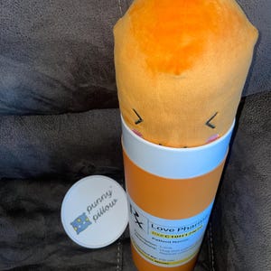 May include: A plush orange pill shaped pillow in a pill bottle container. The bottle is labeled "Love Pharma" and has a prescription number "RX# C10H12N2O".