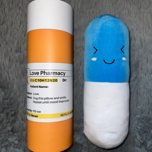 May include: A blue pill-shaped plush toy with a smiling face and a white bottom. The toy is next to a yellow and white cardboard tube with the text "Love Pharmacy" and "RX#:C10H12N20 Dr:" printed on it.