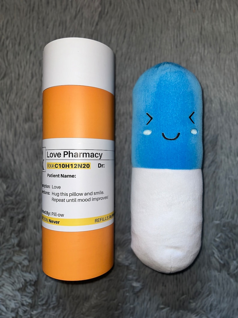 May include: A blue pill-shaped plush toy with a smiling face and a white bottom. The toy is next to a yellow and white cardboard tube with the text "Love Pharmacy" and "RX#:C10H12N20 Dr:" printed on it.