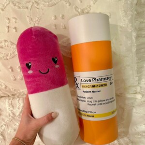 May include: A pink and white pill-shaped plush toy with a smiling face and heart eyes. The toy is next to a tall orange and white container with the text "Love Pharmacy RX#:C10H12N20 Dr. Patient Name: Prescription: Love Directions: Hug this pillow and smile Repeat until mood improves Dispensed By: Pill-ow Refills: Never"