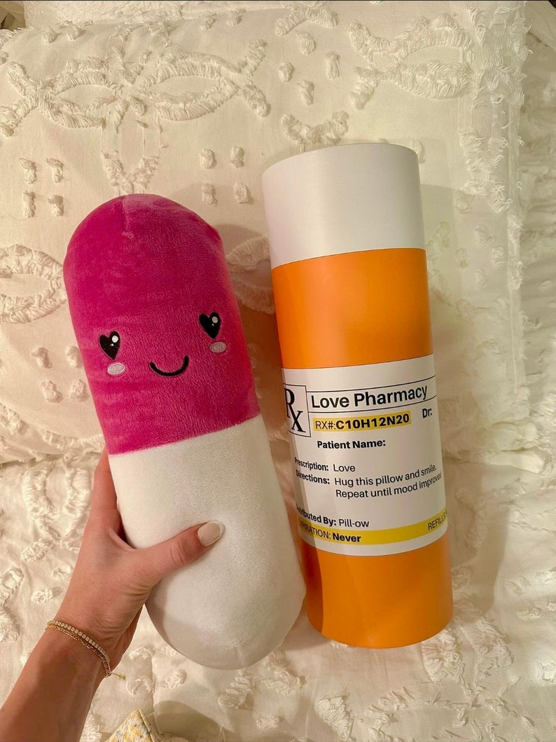 May include: A pink and white pill-shaped plush toy with a smiling face and heart eyes. The toy is next to a tall orange and white container with the text "Love Pharmacy RX#:C10H12N20 Dr. Patient Name: Prescription: Love Directions: Hug this pillow and smile Repeat until mood improves Dispensed By: Pill-ow Refills: Never"
