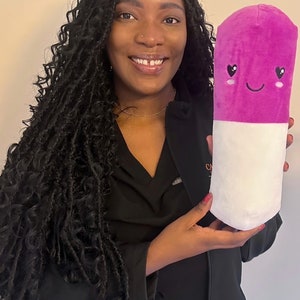 May include: A woman with long black braids is holding a pink and white plush pill with a smiling face and heart eyes.