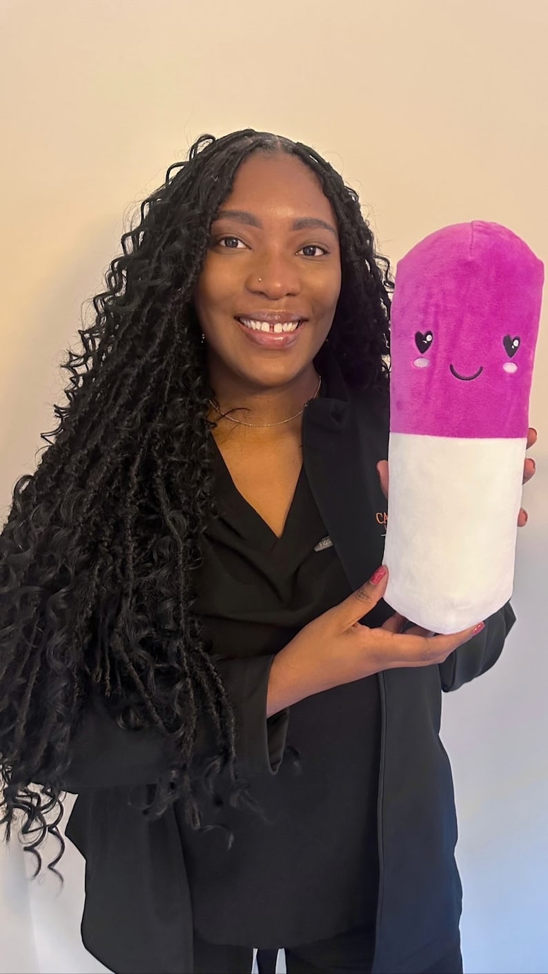May include: A woman with long black braids is holding a pink and white plush pill with a smiling face and heart eyes.