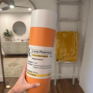 May include: Orange and white pill bottle with the text "Love Pharmacy" and "RX#:C10H12N20 Dr."  The bottle has the text "Prescription: Love" and "Directions: Hug this pillow and smile. Repeat until mood improves."  The bottle also has the text "Distributed By: Pill-ow" and "Expiration: Never".