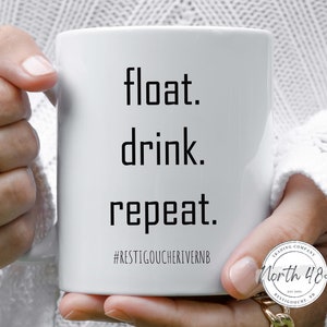 May include: White ceramic mug with the words "float. drink. repeat." in a black, sans-serif font. Below the text is the hashtag #RESTIGOUCHERIVERNB. The mug has a handle and is held by a person.