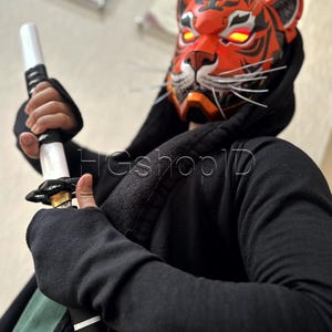 May include: A vibrant orange and black tiger mask with glowing red eyes, paired with a black hooded sweatshirt and a katana. The mask features white whiskers and intricate detailing, creating a striking visual.
