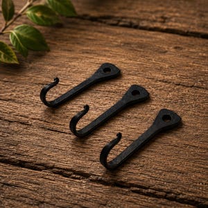 May include: Three black metal hooks with a matte finish, each featuring a curved end and a flat, triangular mounting plate with a single pre-drilled hole at the top for installation. The hooks are arranged in a diagonal row on a wooden surface.