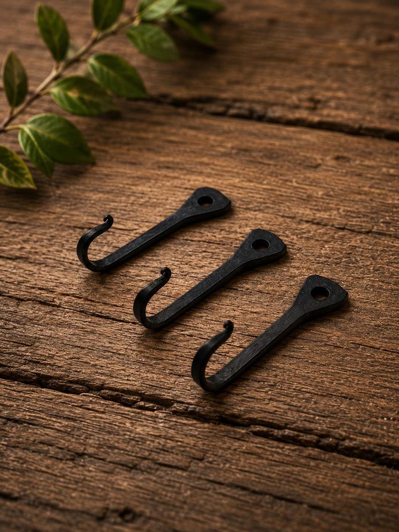 May include: Three black metal hooks with a matte finish, each featuring a curved end and a flat, triangular mounting plate with a single pre-drilled hole at the top for installation. The hooks are arranged in a diagonal row on a wooden surface.
