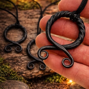 May include: Black metal pendant necklace with a twisted ring and a stylized, symmetrical design. The pendant hangs from a black cord. The background is a rustic wooden surface with moss and soft lighting.