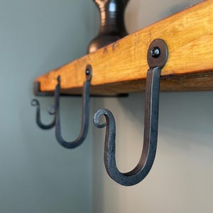 Puede incluir: Tres ganchos de metal negro están unidos a una estantería de madera. Los ganchos son curvos y tienen un remolino decorativo en la parte superior. La estantería está hecha de madera recuperada y tiene un acabado natural.